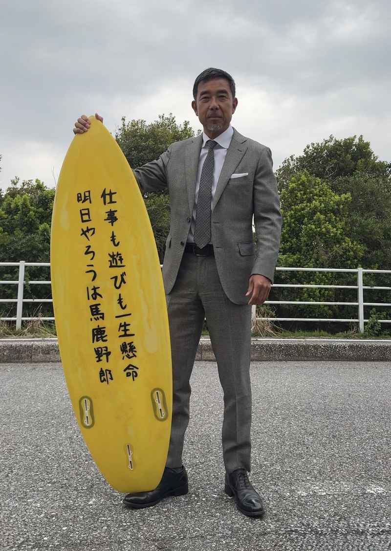 スーツ姿でサーフボードを持つ義忠さん